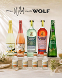 A promotional product lineup of wine and spirits bottles on a draped cream fabric background. Large bottles include a pale yellow wine bottle labeled 'Wild', a pink wine bottle labeled 'Wild Rose', a mint green bottle labeled 'Smiling Wolf Botanique', a red-orange bottle labeled 'Smiling Wolf Apéritif', and a green bottle labeled 'Bottle Green'.